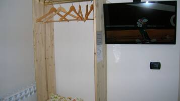 A wooden wardrobe with a hanging rack, a small bed with a floral bedspread, and a television mounted on the wall.