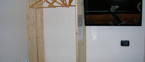A wooden wardrobe with a hanging rack, a small bed with a floral bedspread, and a television mounted on the wall.