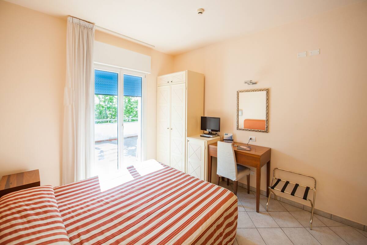 Habitación de hotel con cama, escritorio, computadora, silla, armario y ventana con cortinas.