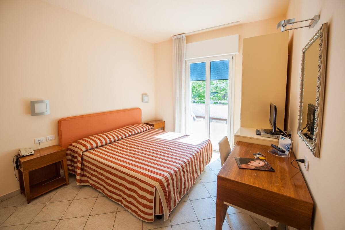 Habitación de hotel con cama, escritorio, espejo y ventana con vistas a zonas verdes.