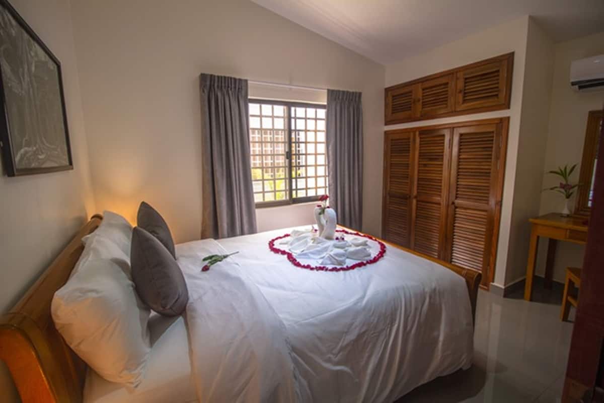 A neatly made bed with a white bedspread and pillows, a window with curtains, and wooden shutters on the wall.