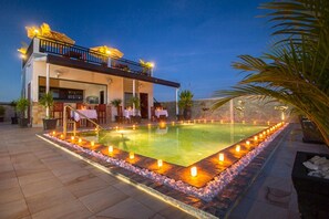 Un espace aménagé au bord de la piscine, avec une piscine, un bâtiment doté d’un balcon et une rangée de bougies disposées le long du bord de la piscine.