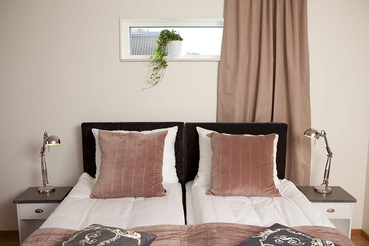 Una cama doble con dos almohadas, una mesita de noche con lámpara, una ventana con planta y una cortina.