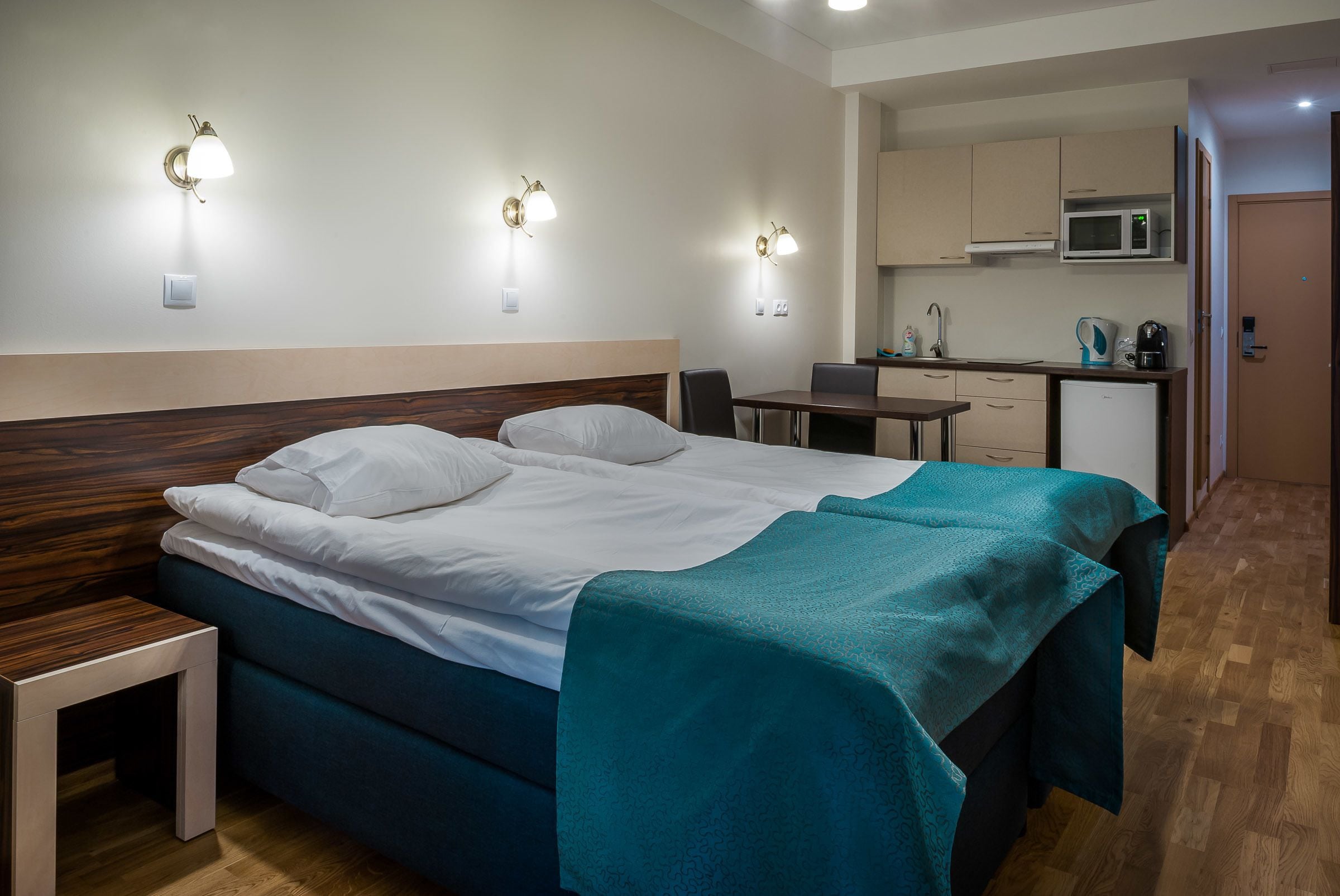 Habitación de hotel con cama, mesita de noche y zona de kitchenette.
