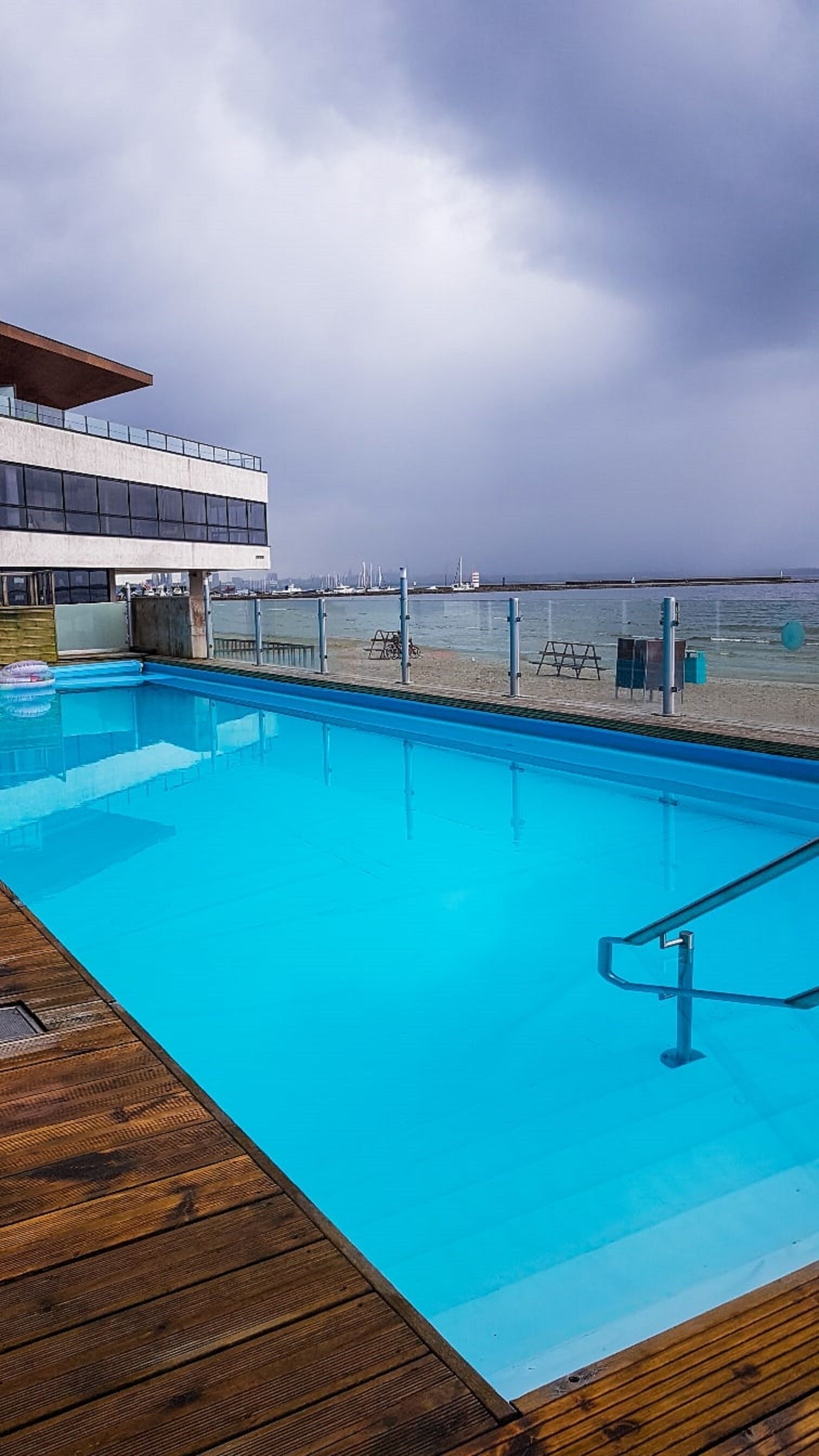 Ein Schwimmbad mit Holzterrasse, ein modernes Gebäude mit großen Fenstern und Blick auf Strand und Meer.