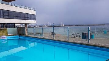 Ein Schwimmbad mit Holzterrasse, ein modernes Gebäude mit großen Fenstern und Blick auf Strand und Meer.