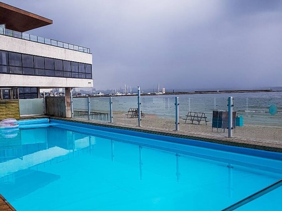 Ein Schwimmbad mit Holzterrasse, ein modernes Gebäude mit großen Fenstern und Blick auf Strand und Meer.