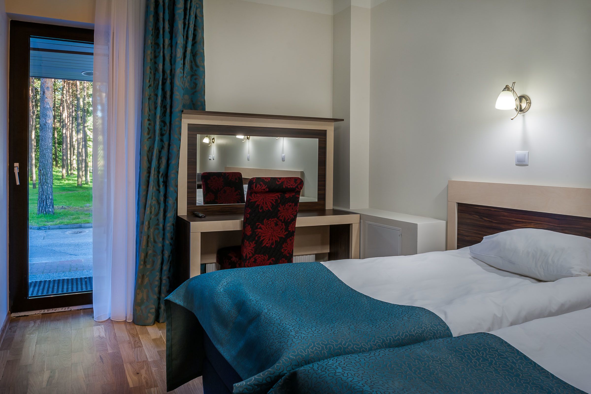 Una camera d'albergo con un letto, una scrivania e una sedia. Si vede una finestra con tende, un comodino e un punto luce a parete.