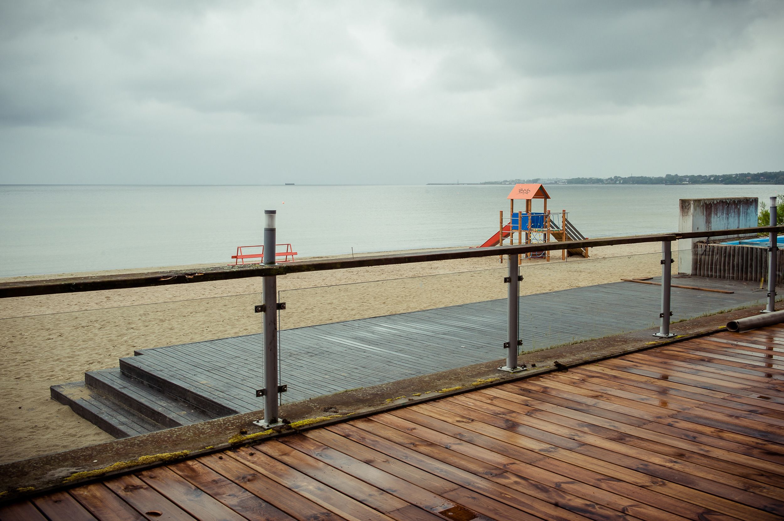 Een houten terras met een metalen hekwerk met uitzicht op een strand met een redderstoren en een glijbaan.