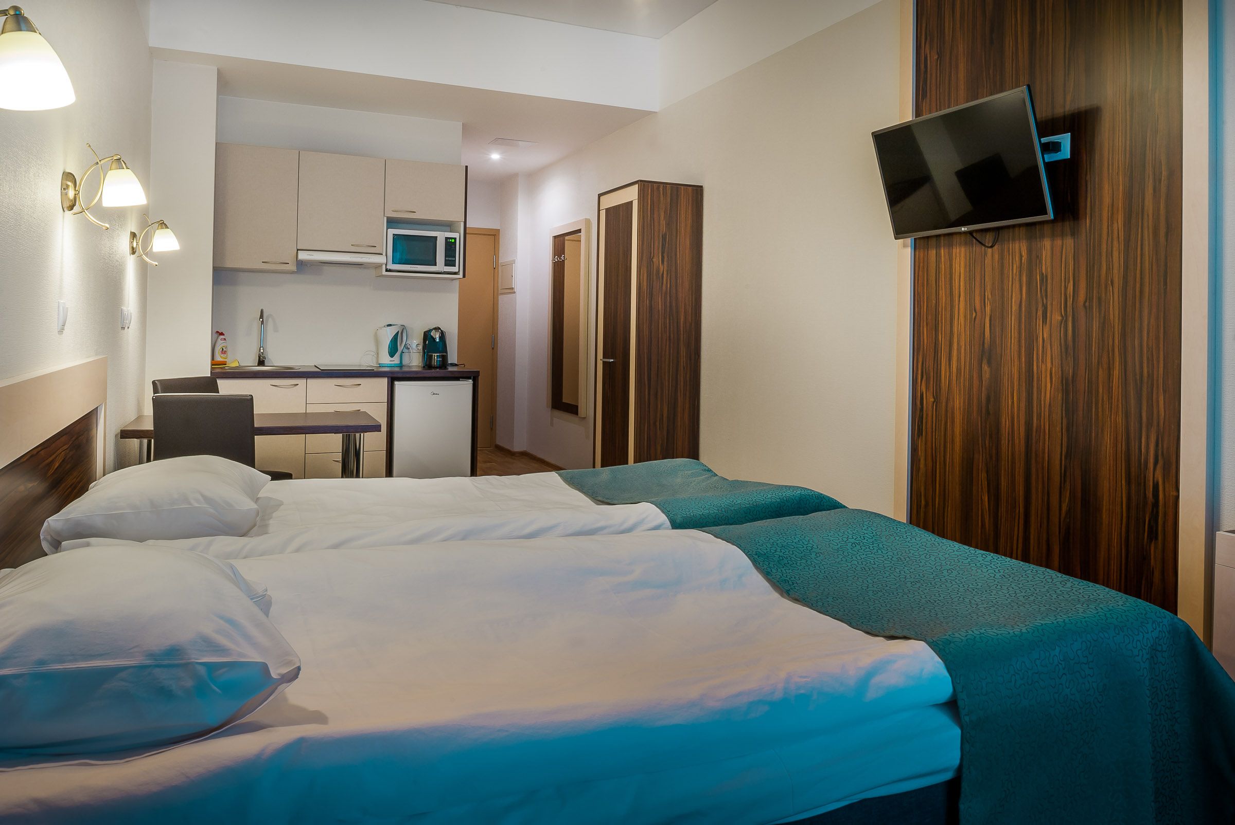 A hotel room with two beds, a TV mounted on the wall, a kitchenette, and a wooden wardrobe.