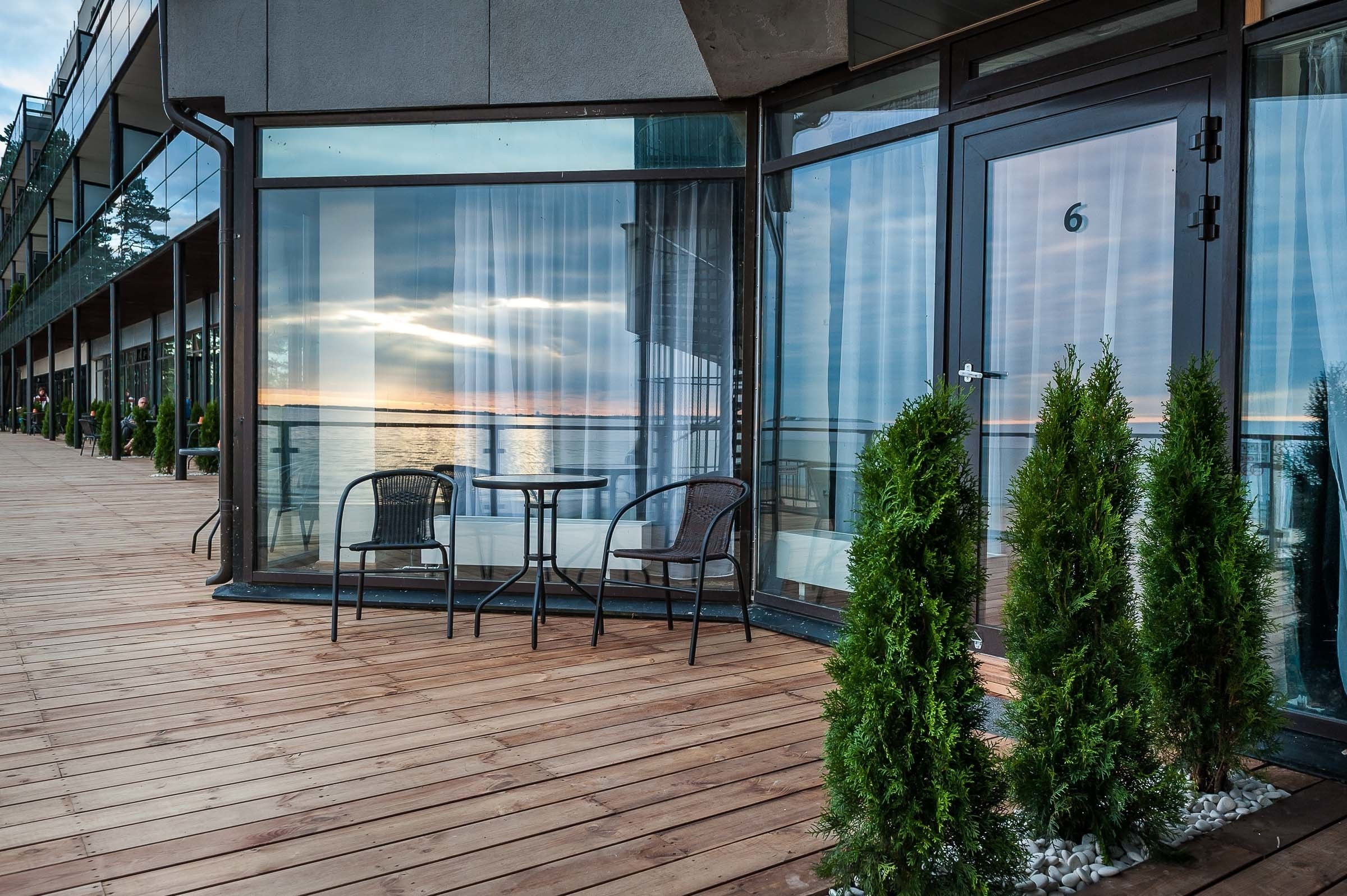 Ein modernes Gebäude mit großen Glasfenstern, einer Holzterrasse mit Sitzgelegenheiten und Blick auf das Wasser.