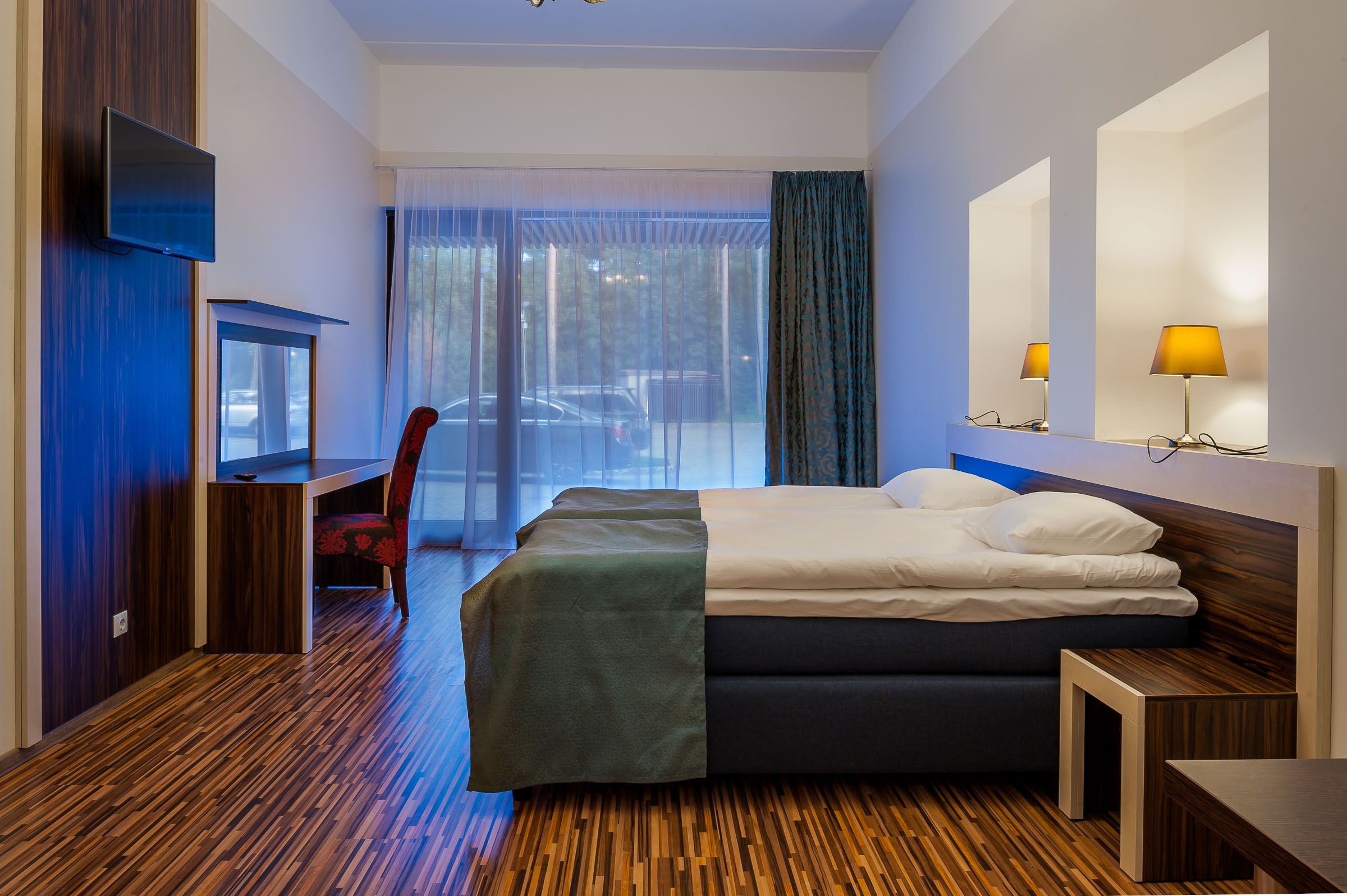 Quarto de hotel moderno com cama, escrivaninha, cadeira, televisão e uma grande janela com cortinas.