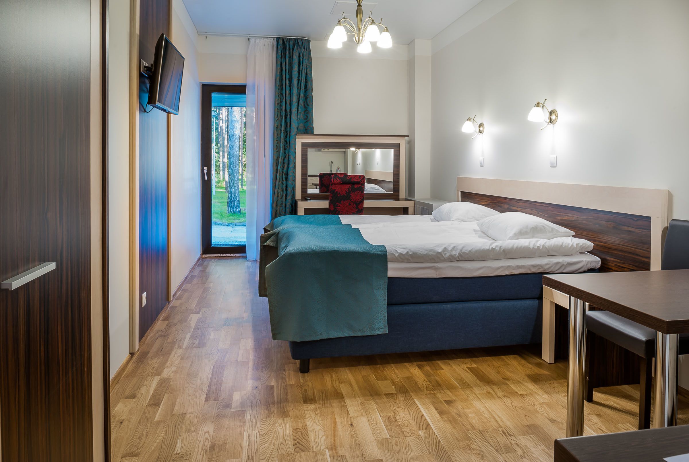 A hotel room with a bed, a desk, a chair, a TV mounted on the wall, and a mirror above the bed.