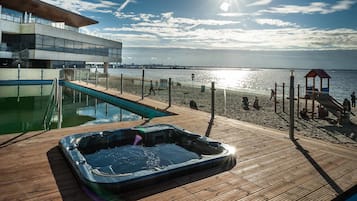 A wooden deck with a hot tub, a swimming pool, and a beach area with a lifeguard tower.