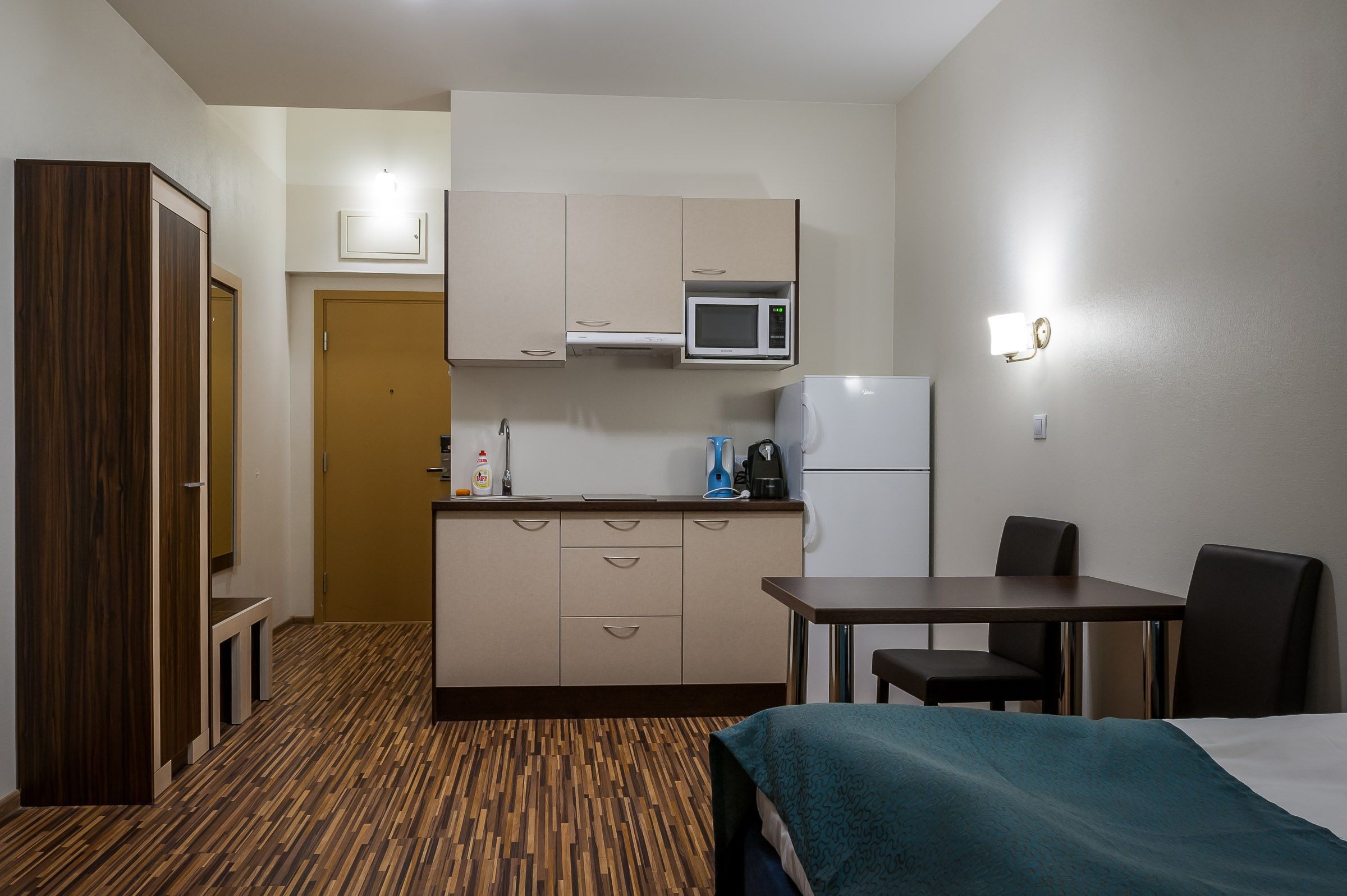 A compact hotel room with a kitchenette, a bed, a small table, and a chair.