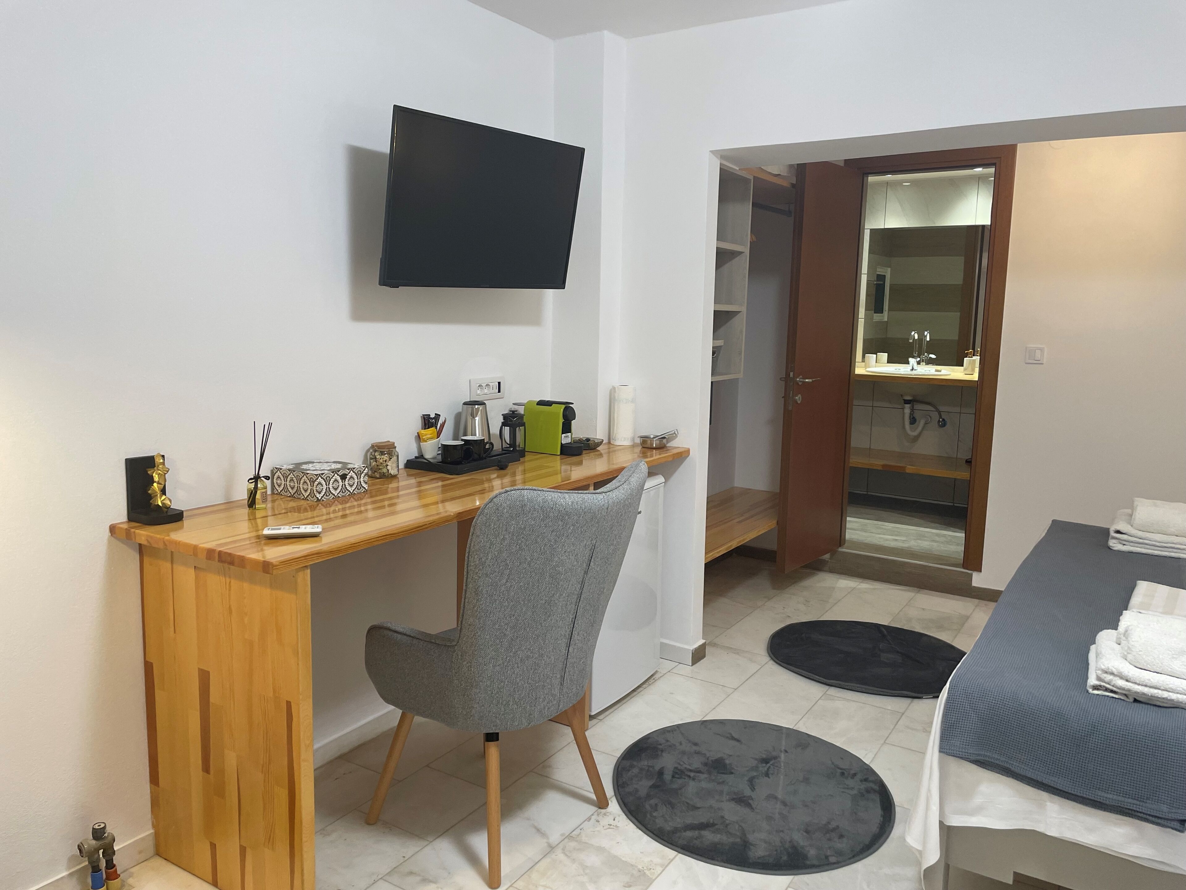 A compact hotel room with a wooden desk, a grey chair, a flat-screen TV, a small kitchenette, and a bathroom visible through an open door.