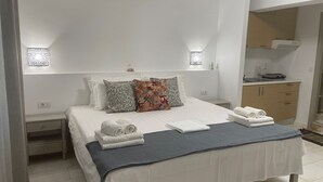 A neatly made bed with white linens, a blue throw blanket, and a decorative pillow. There are two bedside tables with lamps, a small cabinet, and a view of a kitchen area. - Hotel Kourkoumelata (Kefalonia)