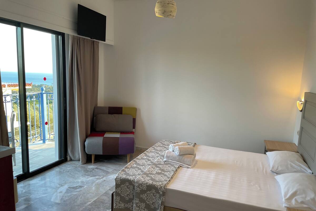 Hotelkamer met een bed, een stoel, een televisie, een balkon met uitzicht en een lamp.