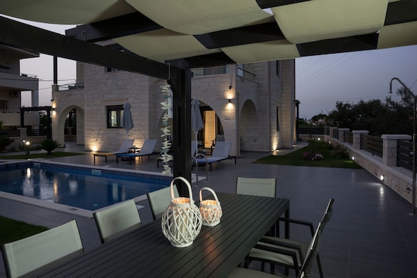 A poolside area with a table, chairs, and lounge chairs. There is a stone building in the background with arched doorways and lighting.