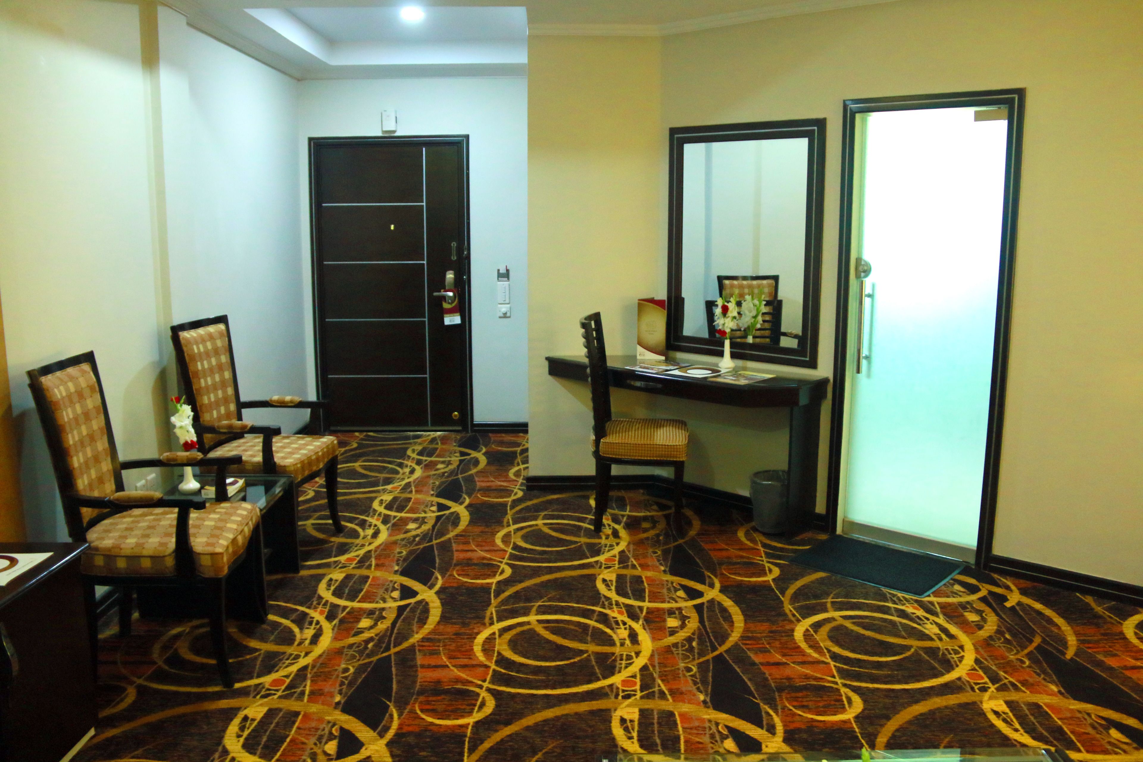 A hotel room with a checkered chair, a desk, a mirror, and a patterned carpet.