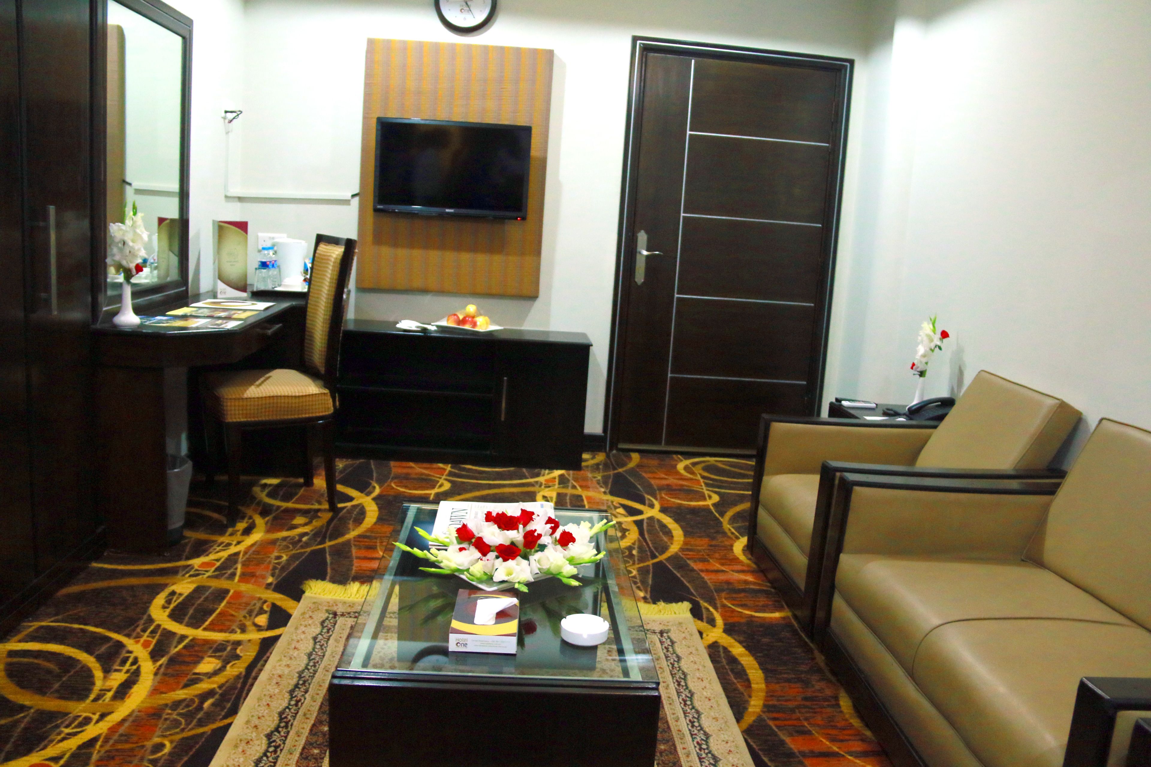 A hotel room with a sofa, a glass coffee table with a floral arrangement, a desk with a chair, a television mounted on the wall, and a wardrobe.