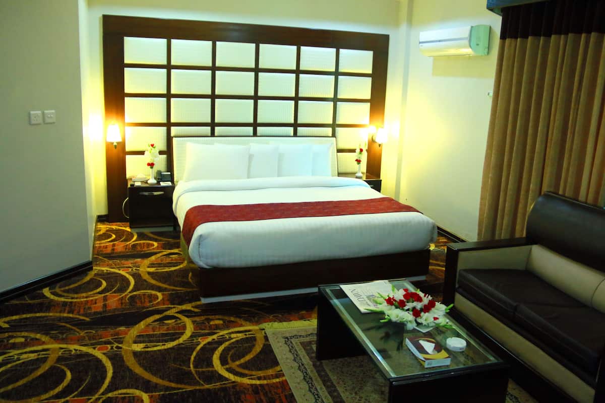 Una habitación de hotel con una cama grande, un sofá, una mesa de centro de vidrio con flores y una mesita de noche con lámpara.