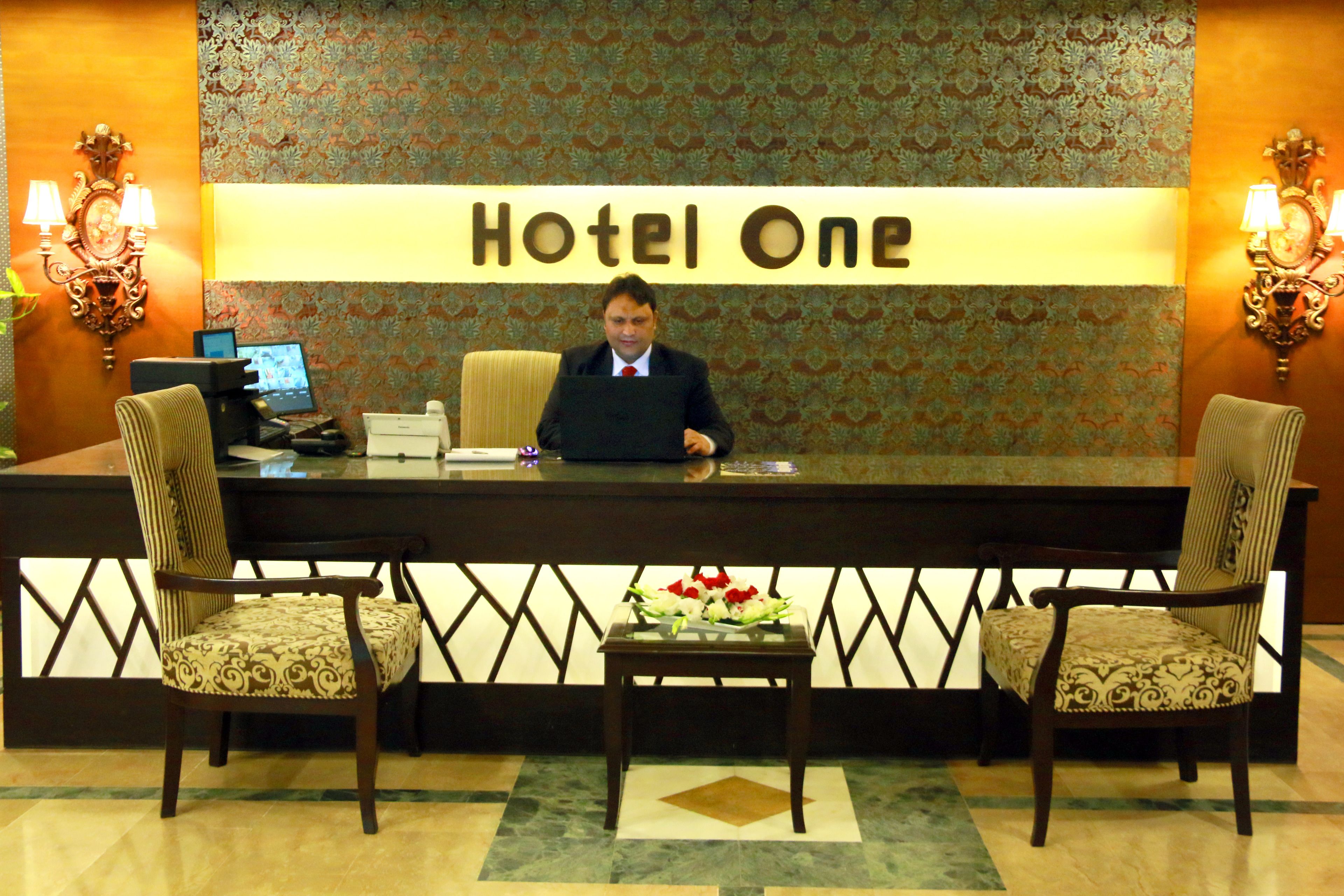 En hotellreception med en skylt som visar texten ”Hotel One”, två stolar, ett litet bord med en blomsteruppsättning och en datorskärm på bordet.