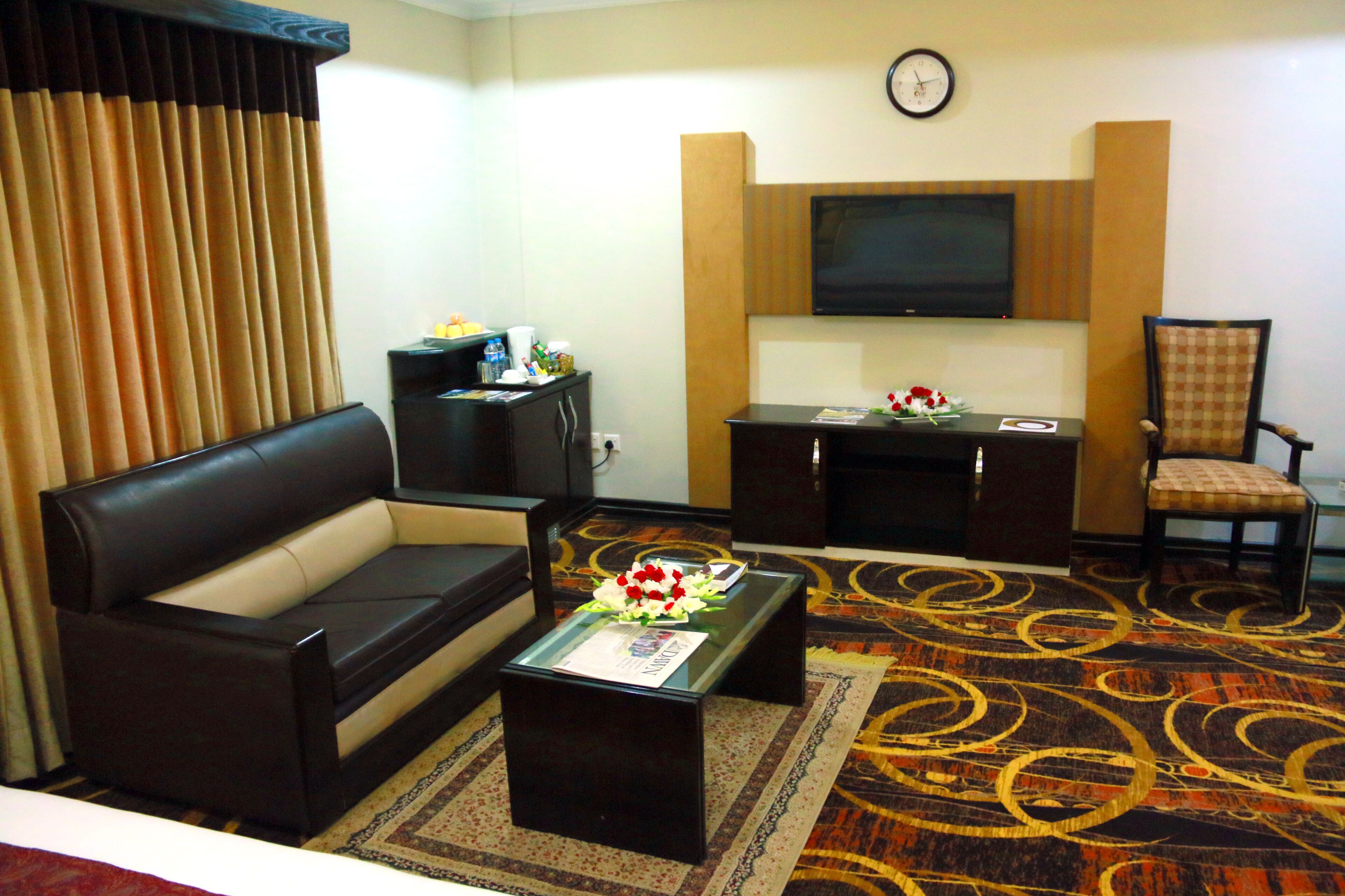 A hotel room with a black leather sofa, a glass coffee table with a flower arrangement, a television mounted on the wall, and a patterned carpet.