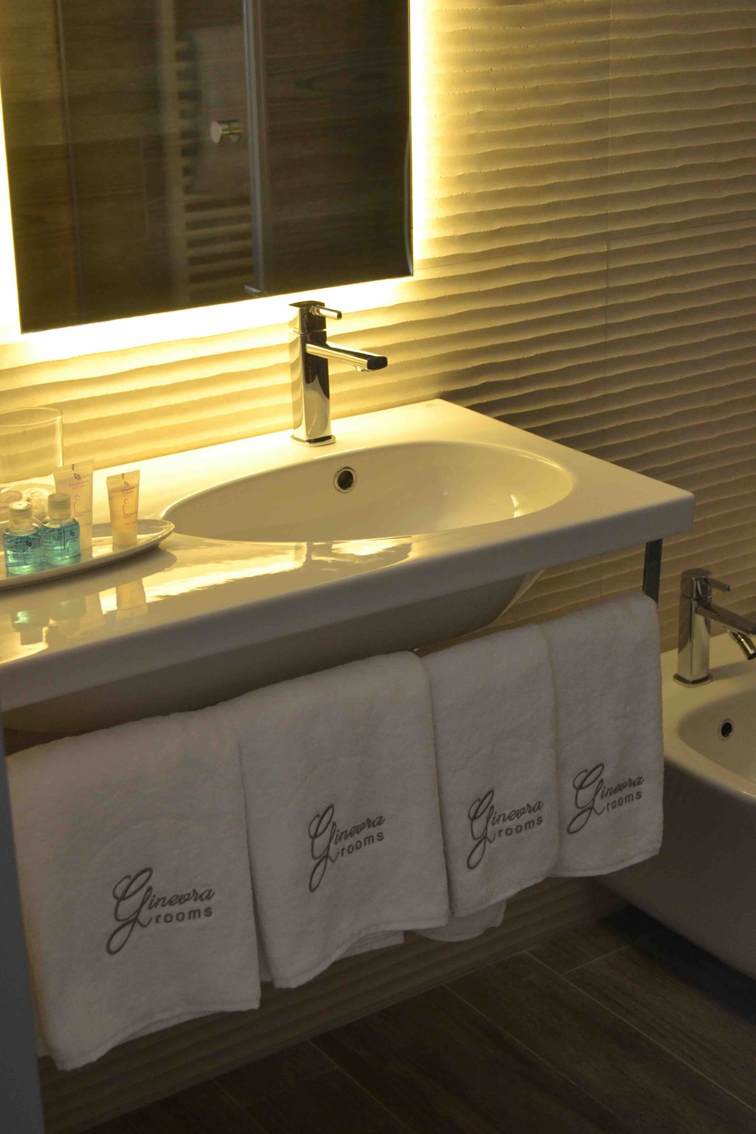 A bathroom with a white sink, a mirror above it, and three towels hanging on a rack labeled "Ginevera rooms."