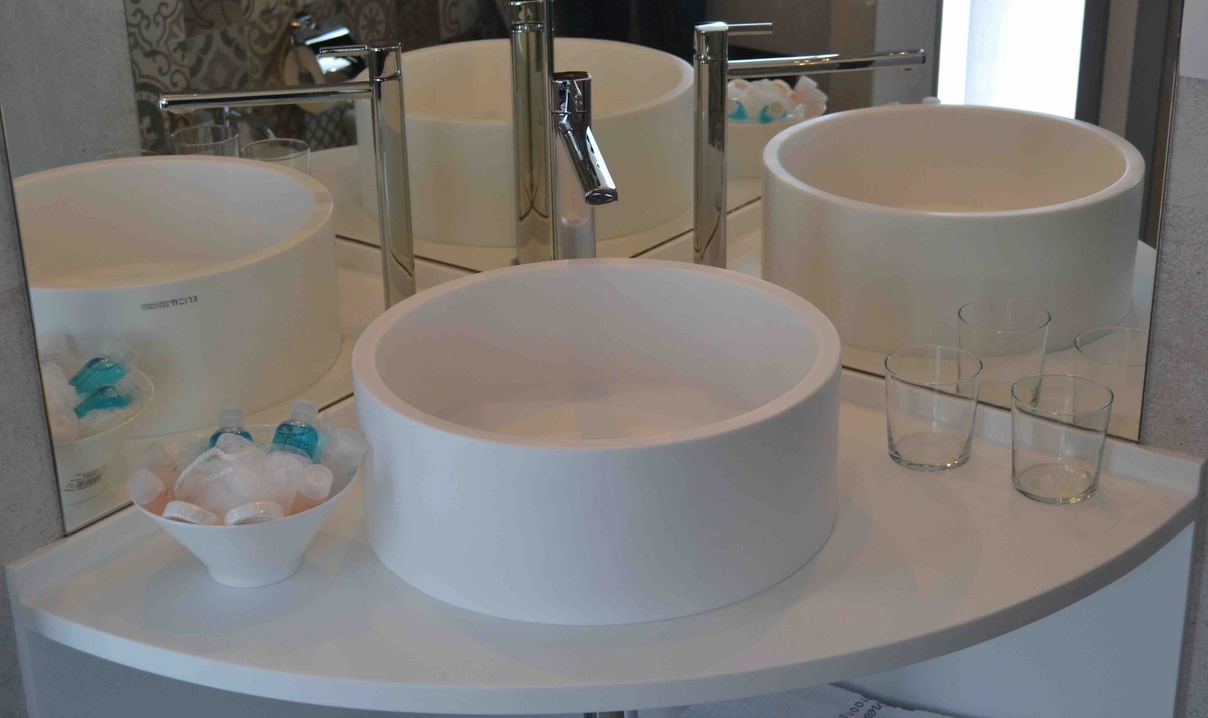 Un baño con tres lavamanos blancos y redondos, un espejo y un recipiente con artículos de aseo.