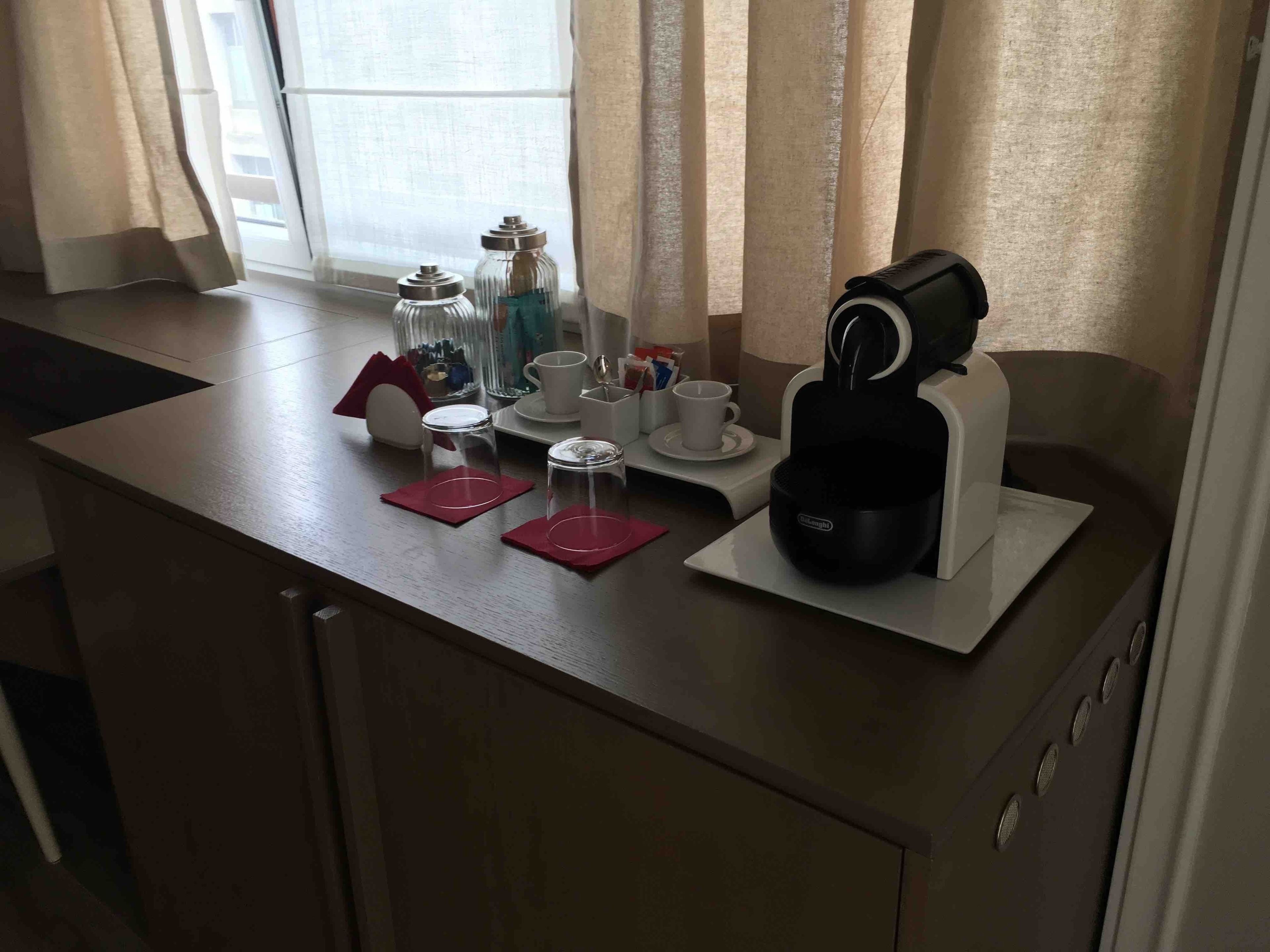 Un taulell de cuina amb una cafetera, dos envasos de vidre, un suport vermell per draps i una tassa blanca.