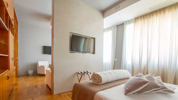 Una habitación de hotel con cama, televisor montado en la pared, armario de madera y ventana con cortinas.