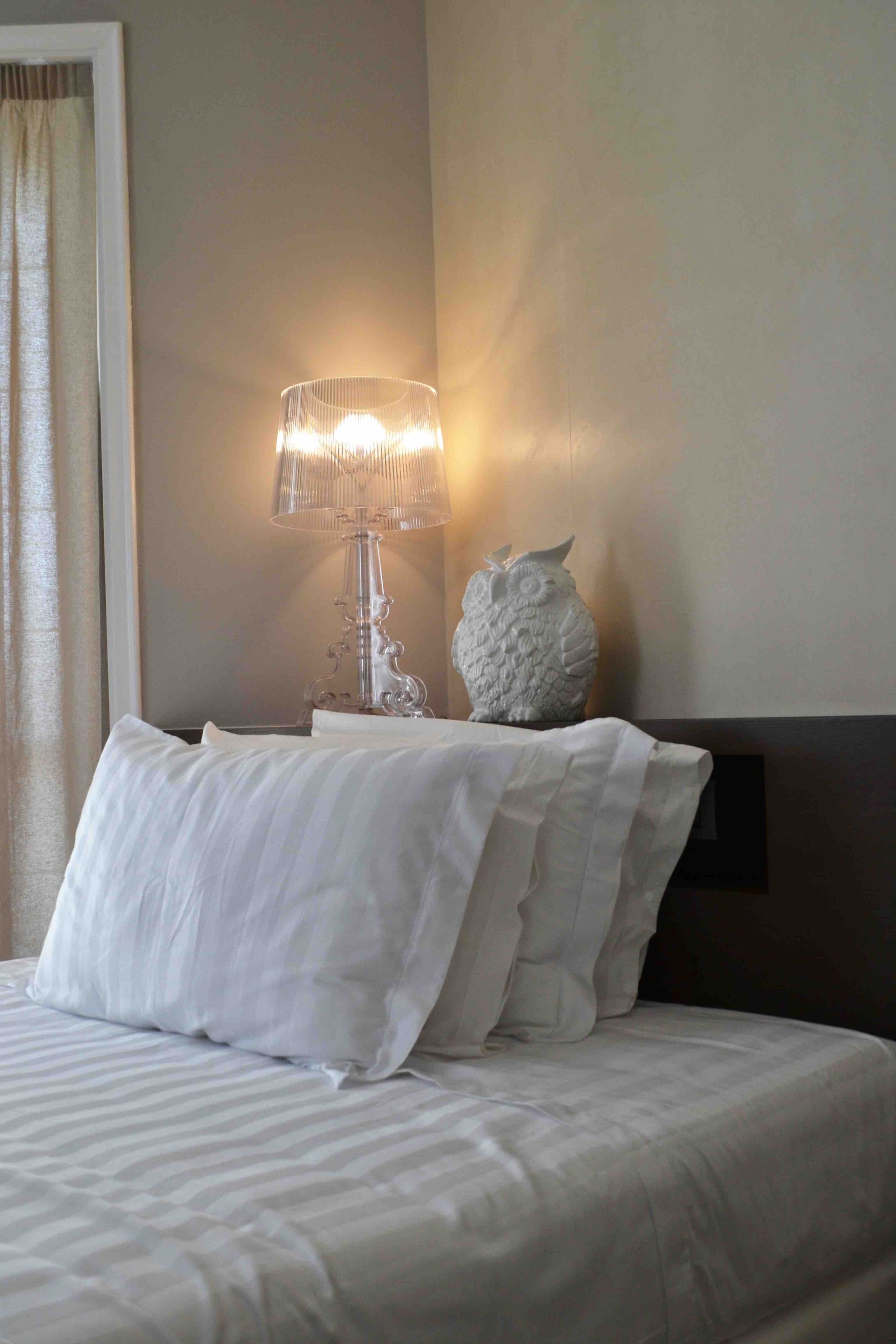 Een opgemaakt bed met witte lakens en kussens, een glazen lamp met meerdere lampenkappen, en een decoratief uiltje op een nachtkastje.