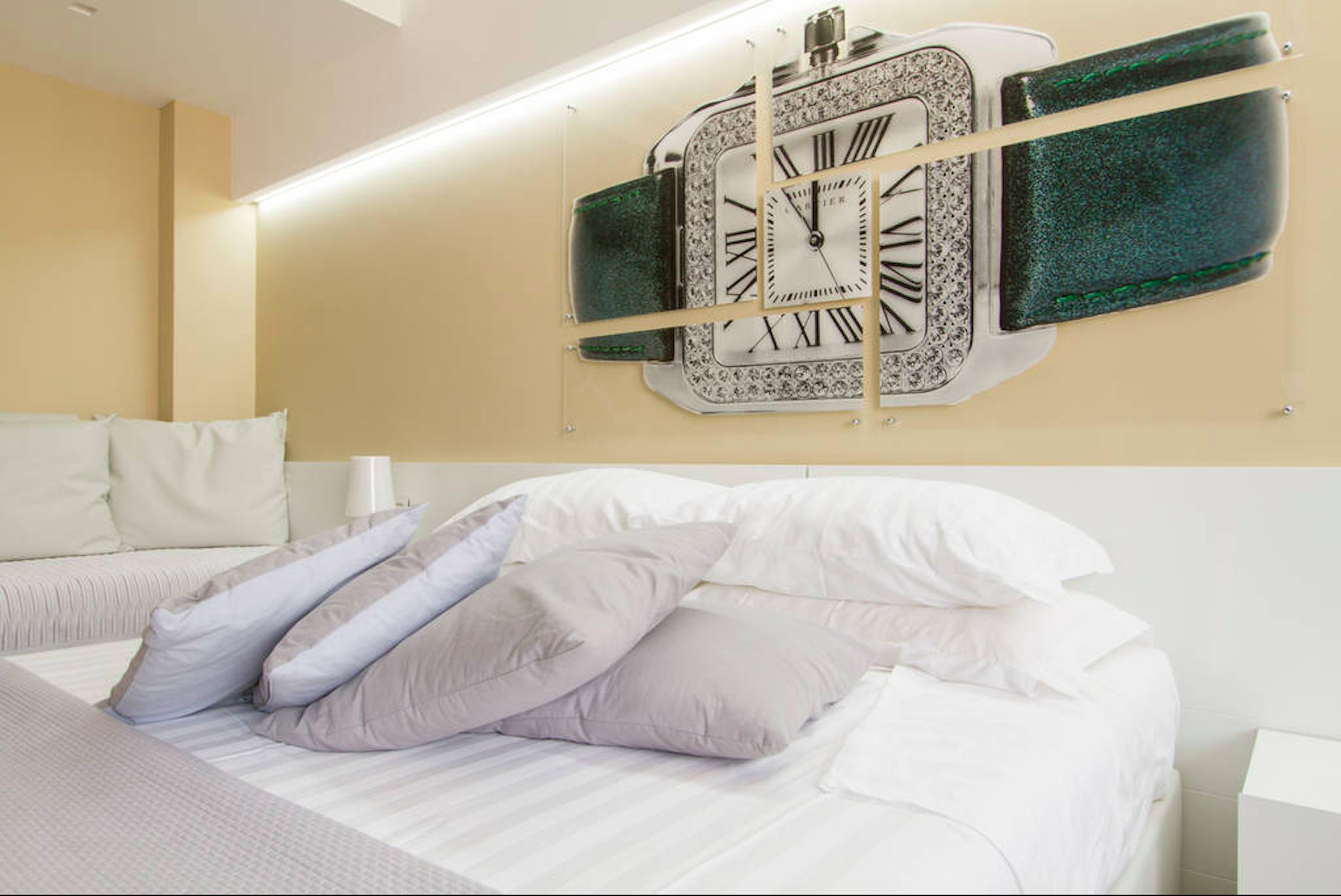 Een hotelkamer met een grote wandklok, een bed met witte beddengoed en meerdere kussens, en een nachtkastje met een lamp.