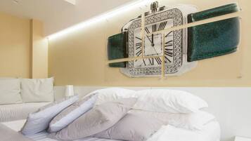 Een hotelkamer met een grote wandklok, een bed met witte beddengoed en meerdere kussens, en een nachtkastje met een lamp.