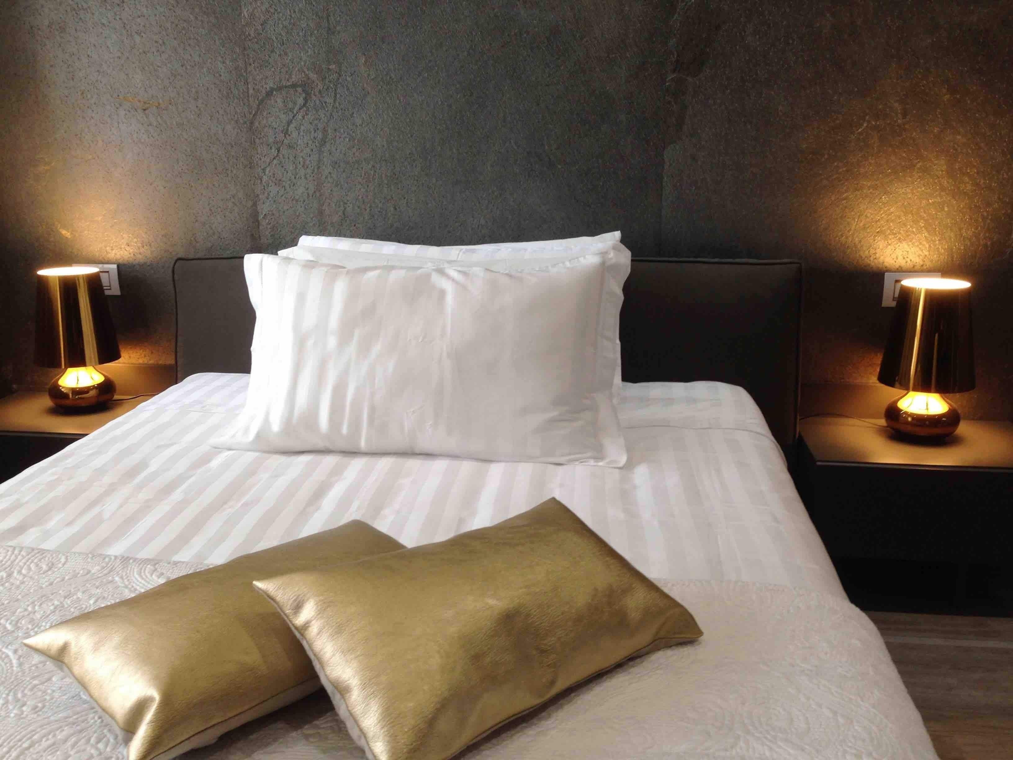 A neatly made bed with white linens and two gold pillows, flanked by two bedside lamps on dark nightstands against a textured wall.