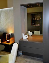 Un bagno moderno con uno specchio grande, una sedia bianca, un tavolino con una lampada e un elemento decorativo a parete.