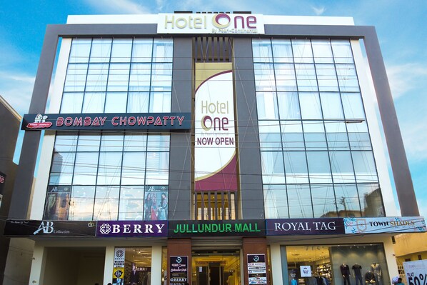 Un hôtel doté d’une grande façade en verre, avec des enseignes indiquant « Hotel One » et « Bombay Chowpatty », ainsi que des devantures de magasins pour « Berry », « Jullundur Mall », « Royal Tag » et « Town Shades ».