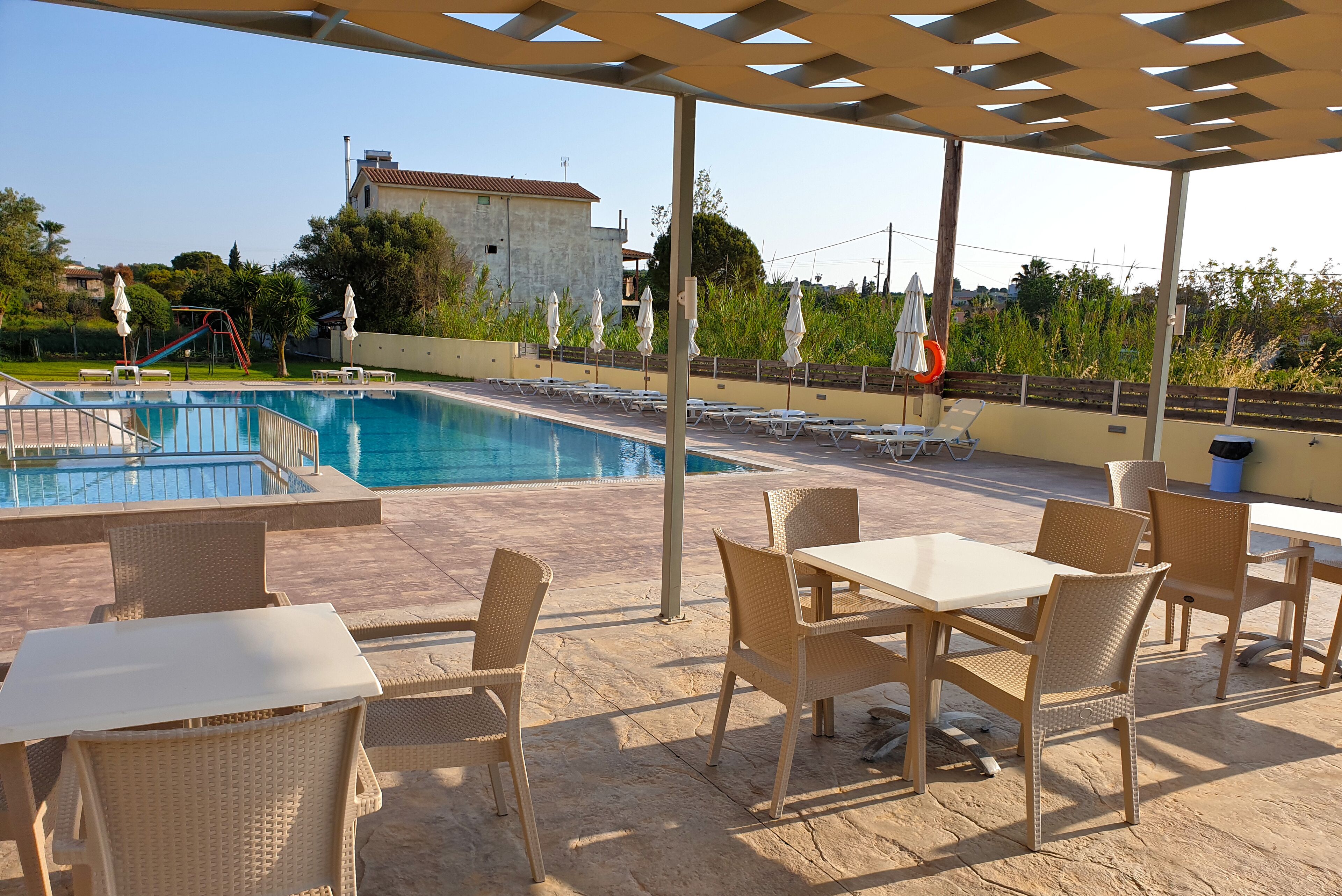Un espace piscine avec des chaises longues et des tables, une terrasse couverte et une vue sur un bâtiment en arrière-plan.