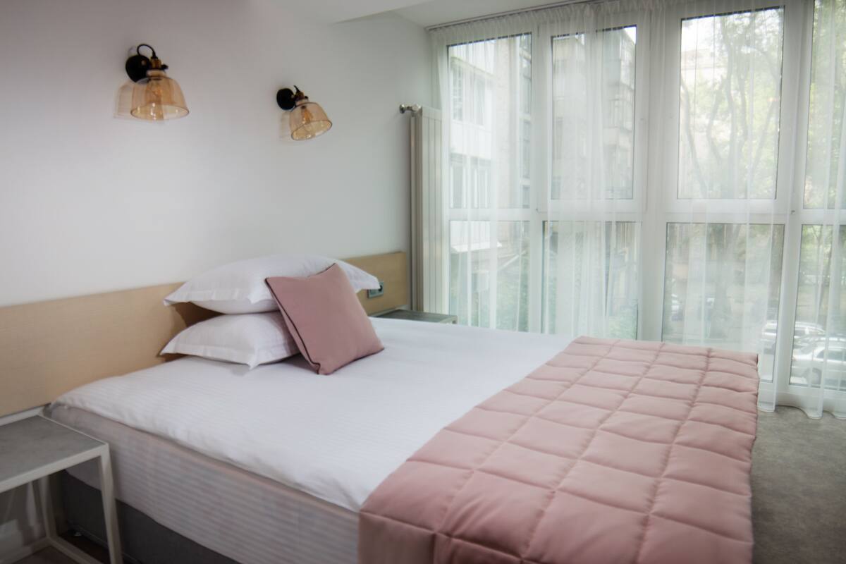 Una cama bien hecha con un edredón rosa, almohadas blancas y una mesita de noche.