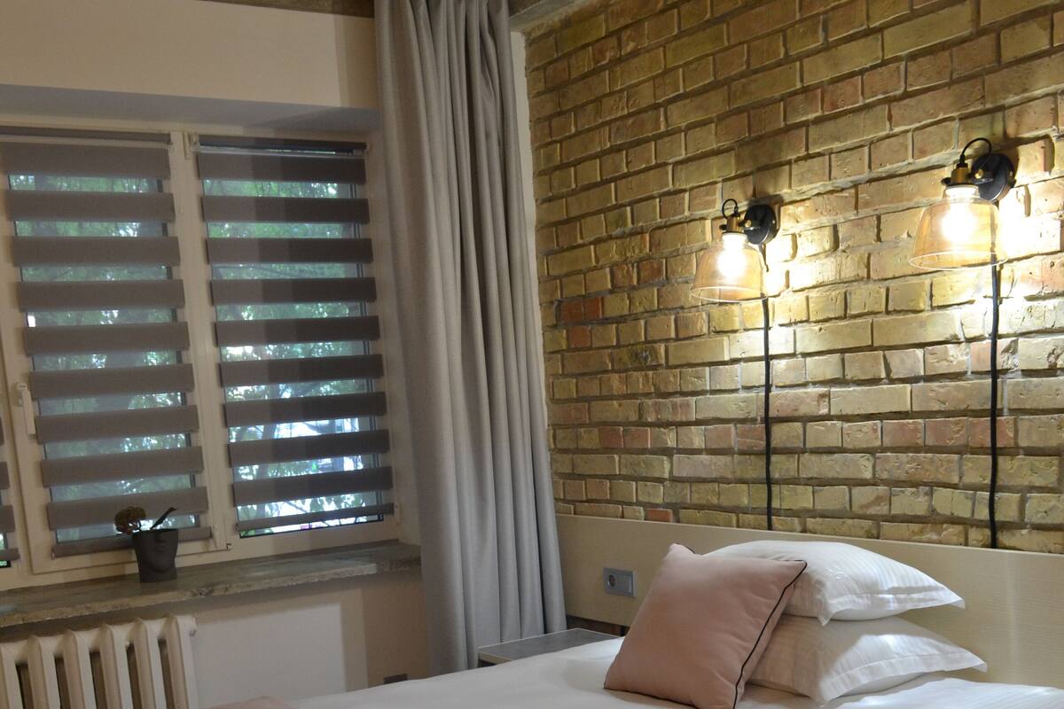 Un dormitorio con pared de ladrillo, una ventana con persianas, una cama con manta rosa y dos luces fijadas a la pared.