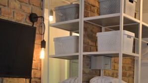 A room with a brick wall, a white shelving unit with baskets, a flat-screen TV, a table with a chair, and a potted plant. - Stan Gret Hotel (Kyiv)