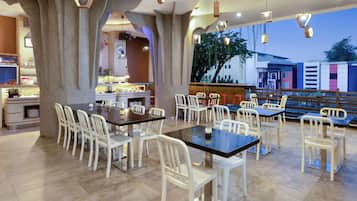Un restaurant au décor moderne, avec une salle à manger centrale dotée de chaises et de tables blanches, et une cuisine visible à travers une arche.