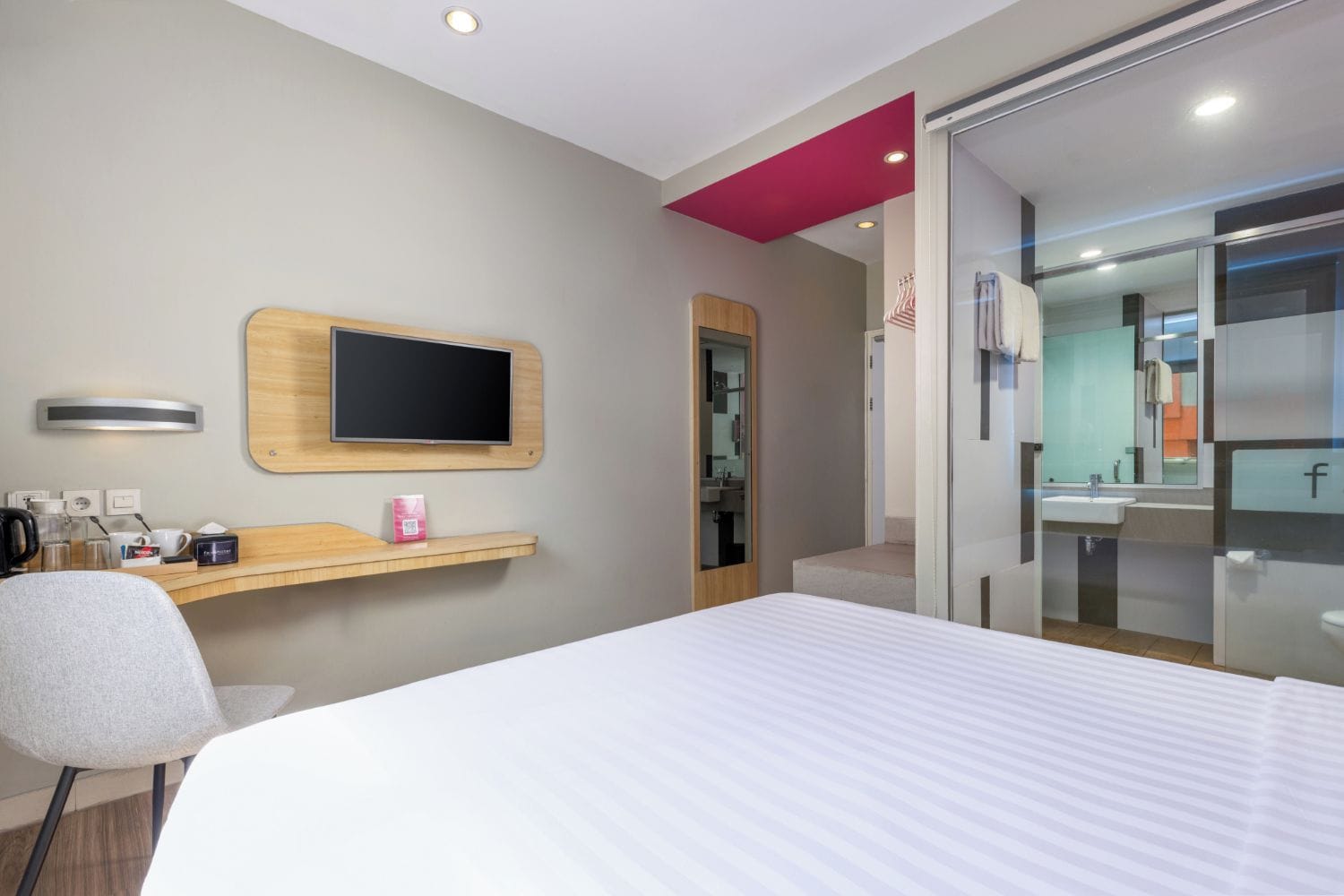 A hotel room with a bed, a desk with a flat-screen TV, a chair, a small shelf with items, and a bathroom with a mirror and sink.