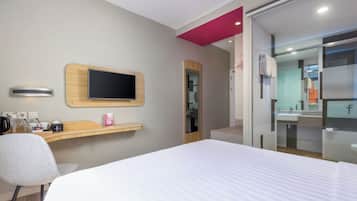 A hotel room with a bed, a desk with a flat-screen TV, a chair, a small shelf with items, and a bathroom with a mirror and sink.