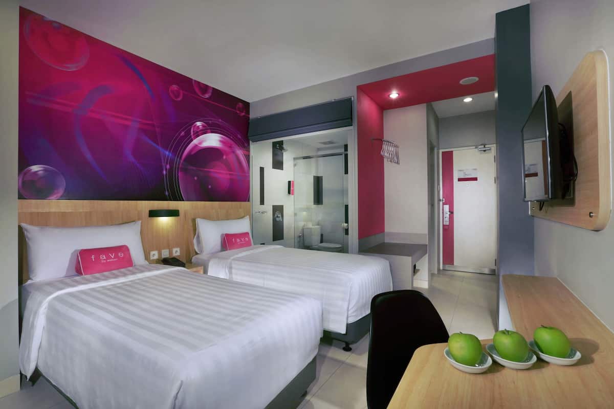 A hotel room with two beds, a desk with apples, a TV, and a bathroom visible through an open door.