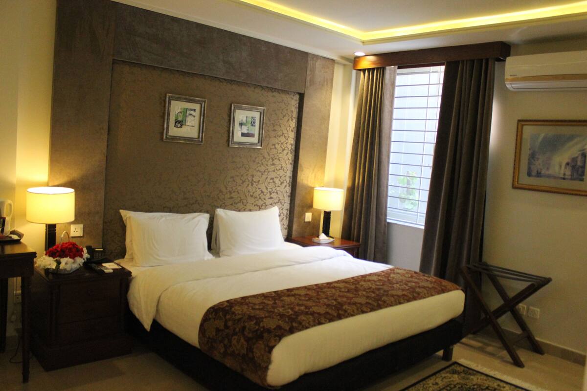 A hotel room with a large bed, two bedside tables with lamps, a small table with a vase of flowers, and a window with curtains.