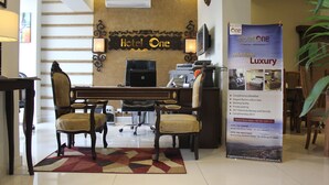 A hotel reception area with a desk, two chairs, a large poster, and a staircase in the background. - Hotel One Super Islamabad (Islamabad)