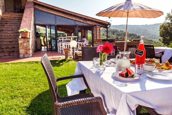 An outdoor dining area with a table set for two, featuring a white tablecloth, a wicker chair, a glass pitcher, a bottle of Succhi Arancia, and plates with food and drinks. - Tenuta Casteani (Gavorrano)