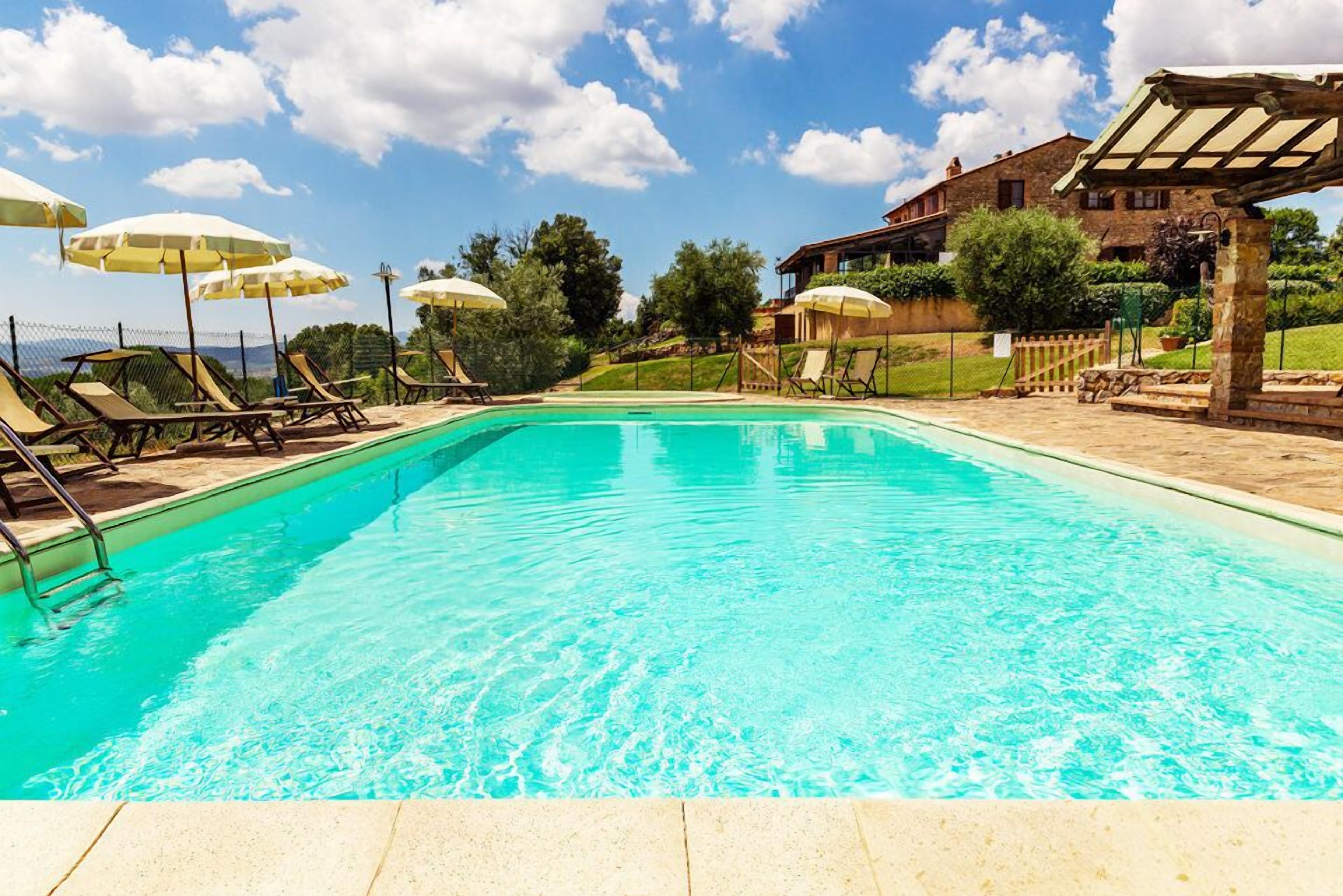 A swimming pool with lounge chairs and umbrellas, surrounded by a stone building and greenery.